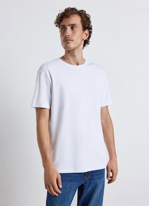 Mann mit lockigem Haar, der ein schlichtes weißes kurzärmeliges T-Shirt und blaue Jeans trägt, blickt leicht seitlich vor einem neutralen Hintergrund.