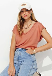 T-shirt rose à col en V avec des manches courtes, associé à un jean en denim léger bleu clair taille haute. Accessoirisé avec une casquette blanche et des colliers en couches.