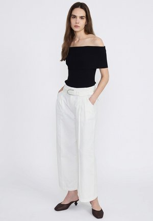 Donna con top nero scollato sulle spalle, pantaloni bianchi a vita alta con cintura, mani nelle tasche e scarpe nere con tacco basso, in piedi su sfondo bianco.