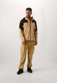 Veste à capuche beige clair avec accents noirs, fermeture éclair et poignets élastiques ; associée à un pantalon cargo beige avec plusieurs poches.