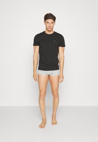Juodas trumpas rankovė t-shirt su maža logotipu ant krūtinės, derinamas su pilkais šortais. Modelis stovi basomis ant vientiso fono.
