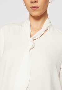 Bílá halenka vyrobená z měkkého materiálu, s uzlem na krku a hladkým, beztexturovým povrchem. Viditelný zlatý přívěsek na náhrdelníku.