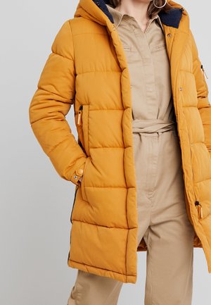 Femme portant un manteau matelassé jaune moutarde avec capuche par-dessus une combinaison beige ceinturée, avec la main dans la poche du manteau.