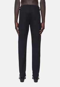 Man som står barfota iförd svarta byxor och svarta lädersandaler, visad från midjan och neråt med avslappnade händer vid sidorna.