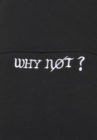 Texte brodé en blanc disant "WHY not ?" avec une ligne diagonale à travers la lettre "o" sur un tissu noir.