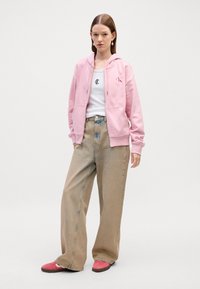Felpa rosa con cappuccio e laccetti, loghi ricamati, t-shirt bianca, pantaloni beige a gamba larga e scarpe rosa con suola testurizzata.