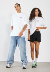 Camisetas blancas de gran tamaño con logo, jeans de tiro bajo color azul claro, pantalones cortos negros con cinturón y zapatillas con cordones verdes y calcetines blancos.