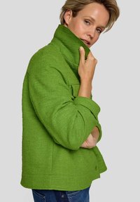 Grüne Strukturjacke mit hohem Kragen, Knopfverschluss und Fronttaschen. Das Modell trägt sie über blauen Jeans und zeigt so die lebendige Farbe.