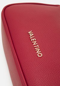 Czerwona skórzana torba o teksturowanej powierzchni, z wytłoczonym złotym logo "VALENTINO" z przodu, w zaokrąglonym kształcie i kontrastującymi wykończeniami.