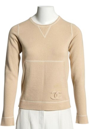 Pull beige à manches longues avec col rond, logo texturé en bas devant, coutures visibles, porté sur une jupe ou un pantalon blanc.