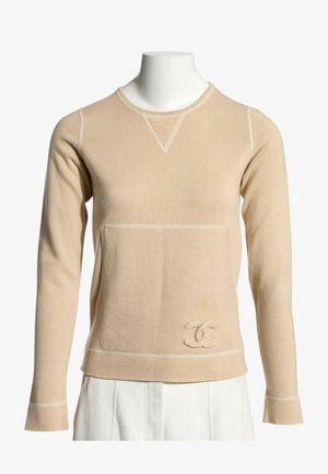 Pull beige à manches longues avec col rond, logo texturé en bas devant, coutures visibles, porté sur une jupe ou un pantalon blanc.