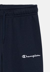 Tums zilas sporta bikses, kas izgatavotas no mīksta kokvilnas sastāva, ar elastīgu jostu un baltu izšūtu "Champion" logotipu kreisajā pusē.