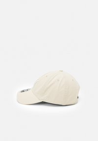 Beige Baumwoll-Baseballmütze mit gebogenem Schirm, besticktem Logo auf der Vorderseite und einem kleinen Textdetail an der Seite.