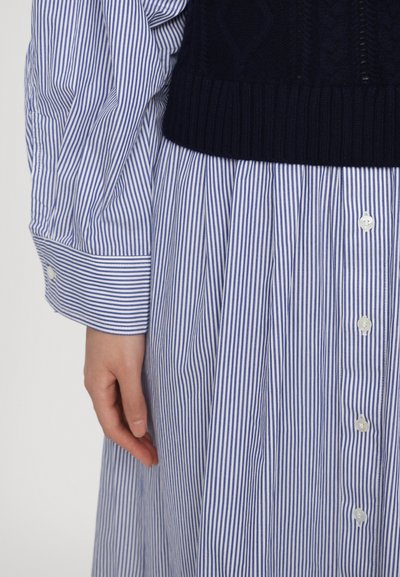Marineblauer Strickpullover, der über einem hellblau-weiß gestreiften Hemdkleid mit Knopfdetails getragen wird, das einen gefältelten Rock und umgeschlagene Ärmel hat.