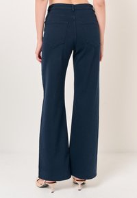 Pantaloni a palazzo blu scuro con vita alta e due tasche posteriori. Realizzati in denim con una consistenza liscia e orli finiti.