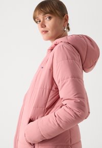 Roze pufferjack met capuchon, gewatteerd ontwerp, zijzakken en klein logodetail op de borst. Soepele textuur, zacht materiaal.