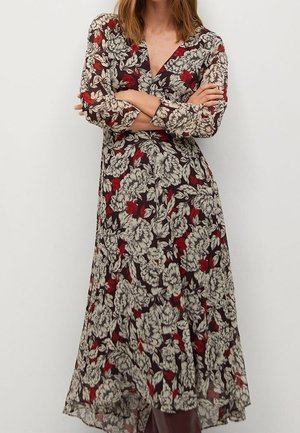 Kvinde iført en langærmet, ankellang kjole med blomsterprint med store hvide blomster og røde accenter på sort baggrund, arme krydset.