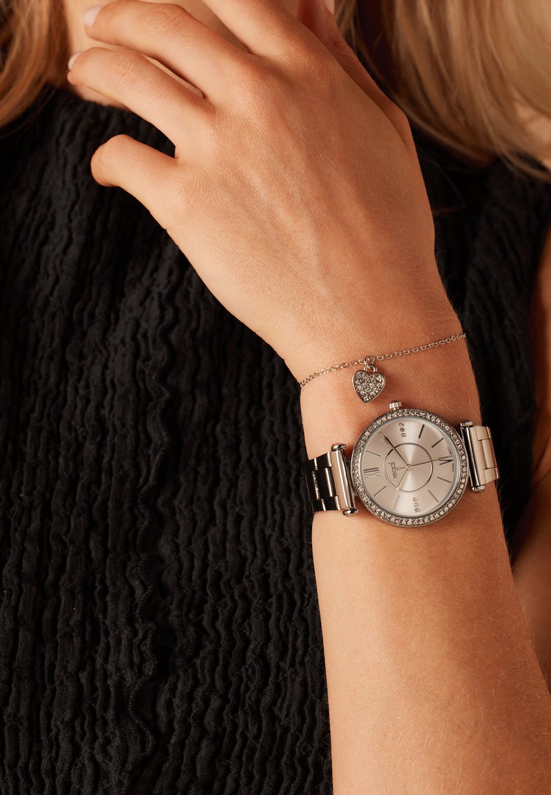 Silberne Uhr mit rundem Ziffernblatt, Kristallakzenten auf der Lünette, Metallarmband, kombiniert mit einem zarten Silberarmband, das einen Herzanhänger trägt.