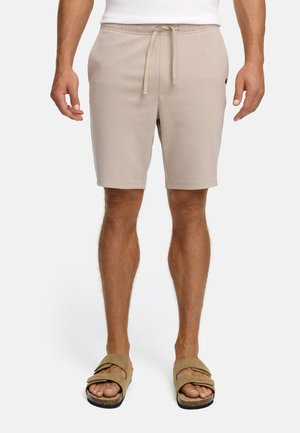 Hombre vistiendo pantalones cortos beige con cordones y sandalias de doble tira color beige, de pie frente a un fondo blanco liso.