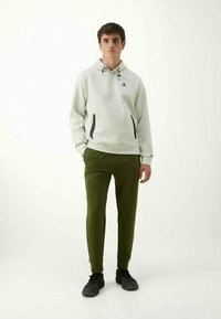 Felpa con cappuccio grigio chiaro con coulisse, dotata di due tasche con zip nere; abbinata a pantaloni verde aderenti e sneakers nere.