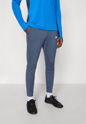 Nike sivi športni hlače s slim fit krojem, stranskimi žepi in belim logotipom; v kombinaciji z modrim dolgim rokavom in črnimi športnimi čevlji.