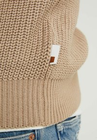 Beige knit sweater with ribbed texture, a small white label featuring a brown square accent on the side, paired with denim.