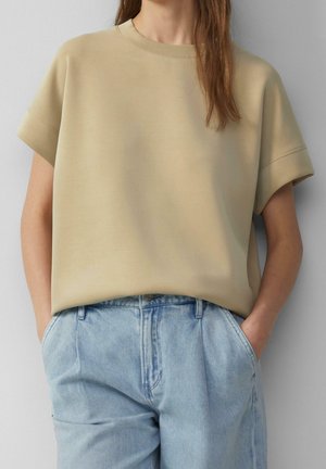 Osoba ubrana w beżową, oversize'ową bluzkę z krótkim rękawem, wpuszczoną w jasnoniebieskie, wysokow-waist jeansy, z rękami w kieszeniach, na szarym tle.