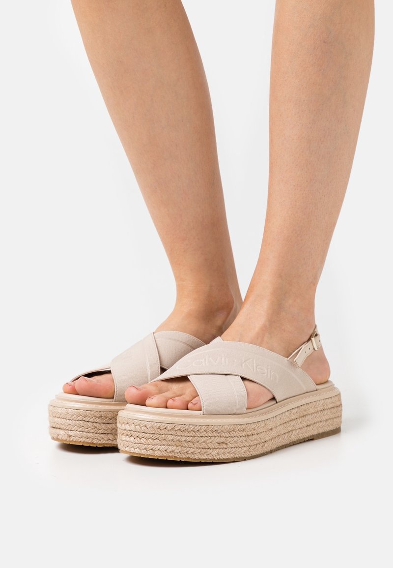 Pieds portant des sandales à plateforme beige avec des brides croisées en tissu et une semelle en corde tressée sur un fond blanc uni.