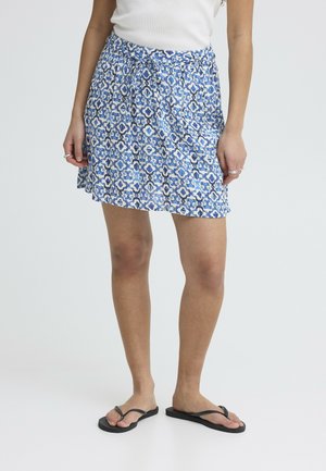 Donna che indossa una gonna a fantasia blu e bianca con cintura annodata, top bianco, anelli argentati e infradito nere su sfondo semplice.