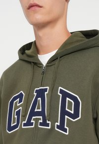 Maslinasto zeleni dukserica sa zipom, sa bijelim i tamnoplavim "GAP" logotipom. Ima vezice i prednji zip, izrađena je od mekanog pamučnog materijala.