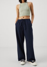 Person som bär en ljusgrön ärmlös kort topp, marinblå vida byxor med dragsko och svarta och vita sneakers, står med händerna i fickorna.