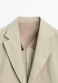 Beige colbert met brede inkeping revers en een gladde binnenvoering, platliggend weergegeven tegen een witte achtergrond.