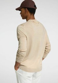 Μπεζ πουλόβερ από πλέξη με μακριά μανίκια, ριμπ μανσέτες και τελείωμα, με στρογγυλή λαιμόκοψη. Το μοντέλο φοράει ανοιχτόχρωμο παντελόνι και καφέ καπέλο.