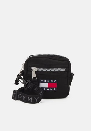 Small black crossbody bag with front zipper pocket, Tommy Jeans logo patch, and adjustable strap with "Tommy" text.