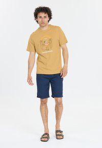 Beige t-shirt med en flamingo-grafik och palmer, i kombination med mörkblå shorts. Shorts är knälånga med uppvikta muddar. Bruna sandaler.