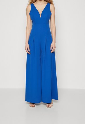Vrouw draagt een mouwloze, koningsblauwe jumpsuit met brede pijpen, een diepe V-hals en geplooide voorkant, gecombineerd met zilveren open teen hakken.