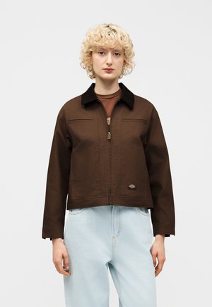 Fiatal személy göndör szőke hajjal, barna cipzáras dzsekit visel fekete gallérral és világoskék farmert, egy egyszerű háttér előtt áll.