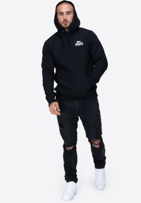 Czarna hoodie z kapturem, z przednią kieszenią kangurką, noszona na zniszczonych czarnych dżinsach i białych sneakersach. Prosty detal z logo na klatce piersiowej.