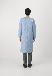 Lång vadderad kappa i ljusblått med diamantmönster, rund halsringning och logotyp på övre delen av ryggen, matchad med grå byxor och svarta skor.