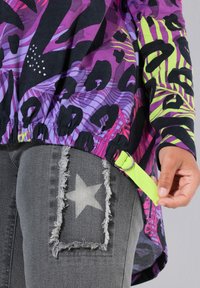 Persona ajustando una correa de color verde neón en una blusa de manga larga con un colorido patrón púrpura y negro, llevando jeans grises con un parche de estrella deshilachado.