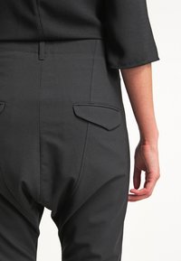 Černé přizpůsobené kalhoty s hladkým materiálem, úzkým střihem a dvěma zadními kapsami s klopami. Pás je strukturovaný pro elegantní vzhled.