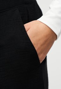 Χέρι με λευκό μανίκι που ξεκουράζεται μέσα στην τσέπη μαύρου παντελονιού με υφή, σε απλό φόντο.