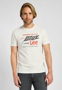 Camiseta de algodón color crema con mangas cortas, que presenta un estampado gráfico en rojo y negro. El texto incluye "UNION ALLS" y "Lee American Original."