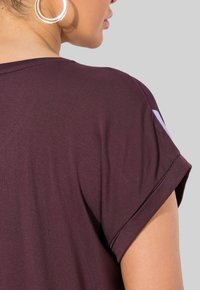 Burgundské tričko s ohrnutými rukávy, měkká textura látky a jemný lesk. Detailní pohled na zadní část ramene, žádné viditelné vzory.