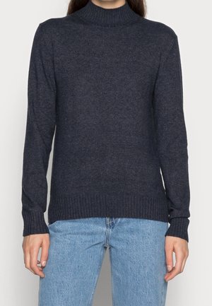 Personne portant un pull à col roulé côtelé bleu marine foncé et un jean en denim bleu clair, montré des épaules jusqu'aux hanches.