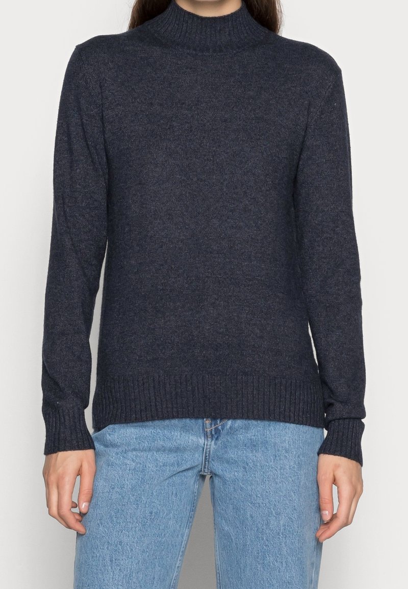 Personne portant un pull à col roulé côtelé bleu marine foncé et un jean en denim bleu clair, montré des épaules jusqu'aux hanches.