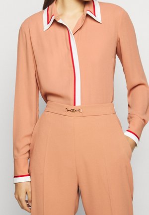 Blusa color pesca con colletto bianco e strisce rosse, maniche lunghe e decorazioni in tonalità oro in vita, caratterizzata da una texture liscia.