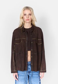 Brun mockaläder skjorta med två bröstfickor, prydd med guldfärgad nitar längs sömmarna. Har en knappstängning framtill och en avslappnad passform.