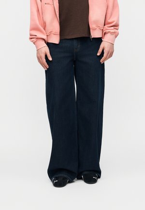 Άτομο που φοράει σκούρο τζιν με φαρδιά πόδια, μαύρα sneakers Puma, καφέ πουκάμισο και ροζ φούτερ με φερμουάρ, στέκεται μπροστά σε απλό φόντο.