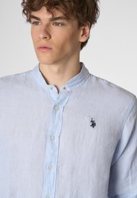 Giovane uomo con capelli ricci che indossa una camicia button-up azzurro chiaro con un piccolo logo scuro sul petto, in piedi contro uno sfondo neutro.